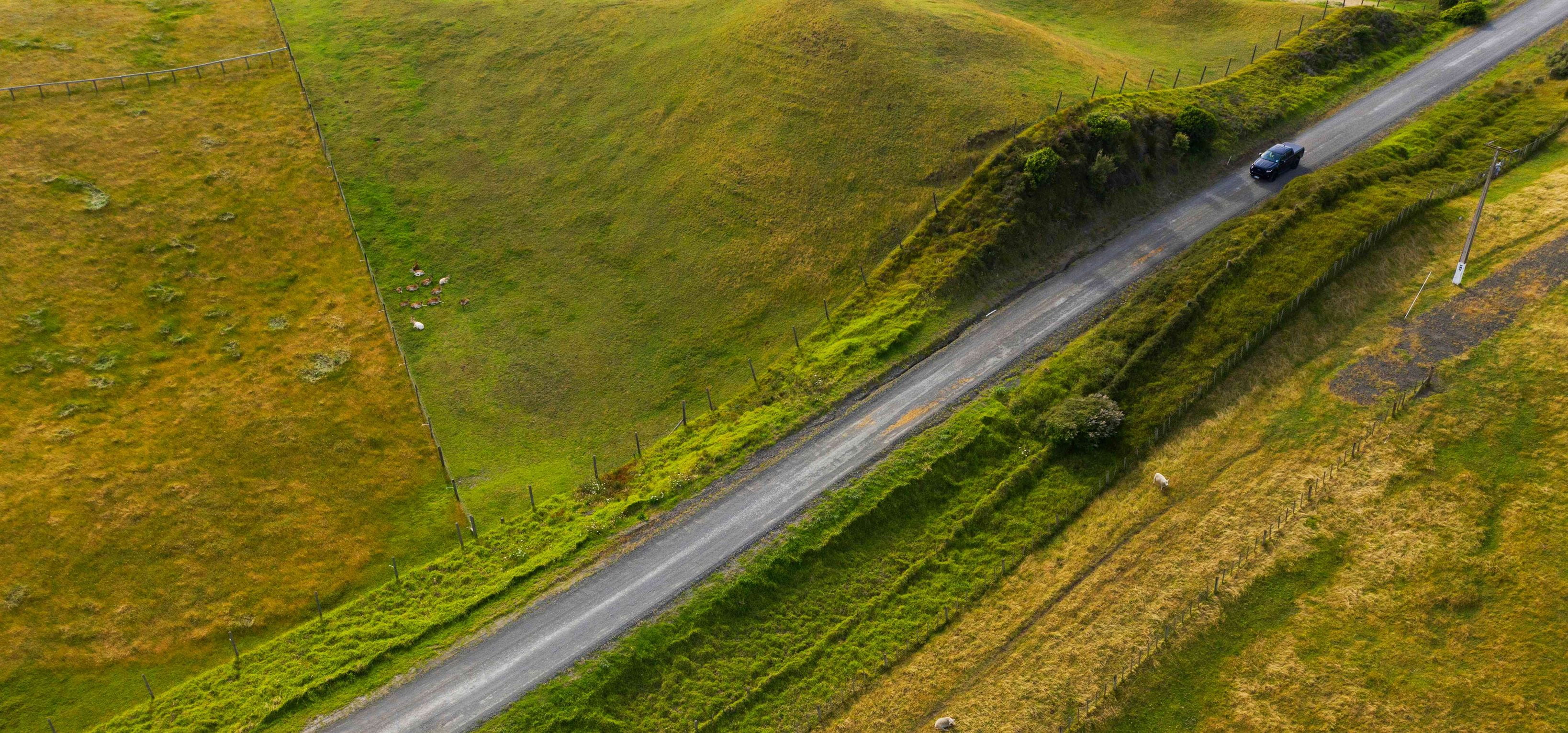 Aerial shot of a black car on a country road surrounded by green fields